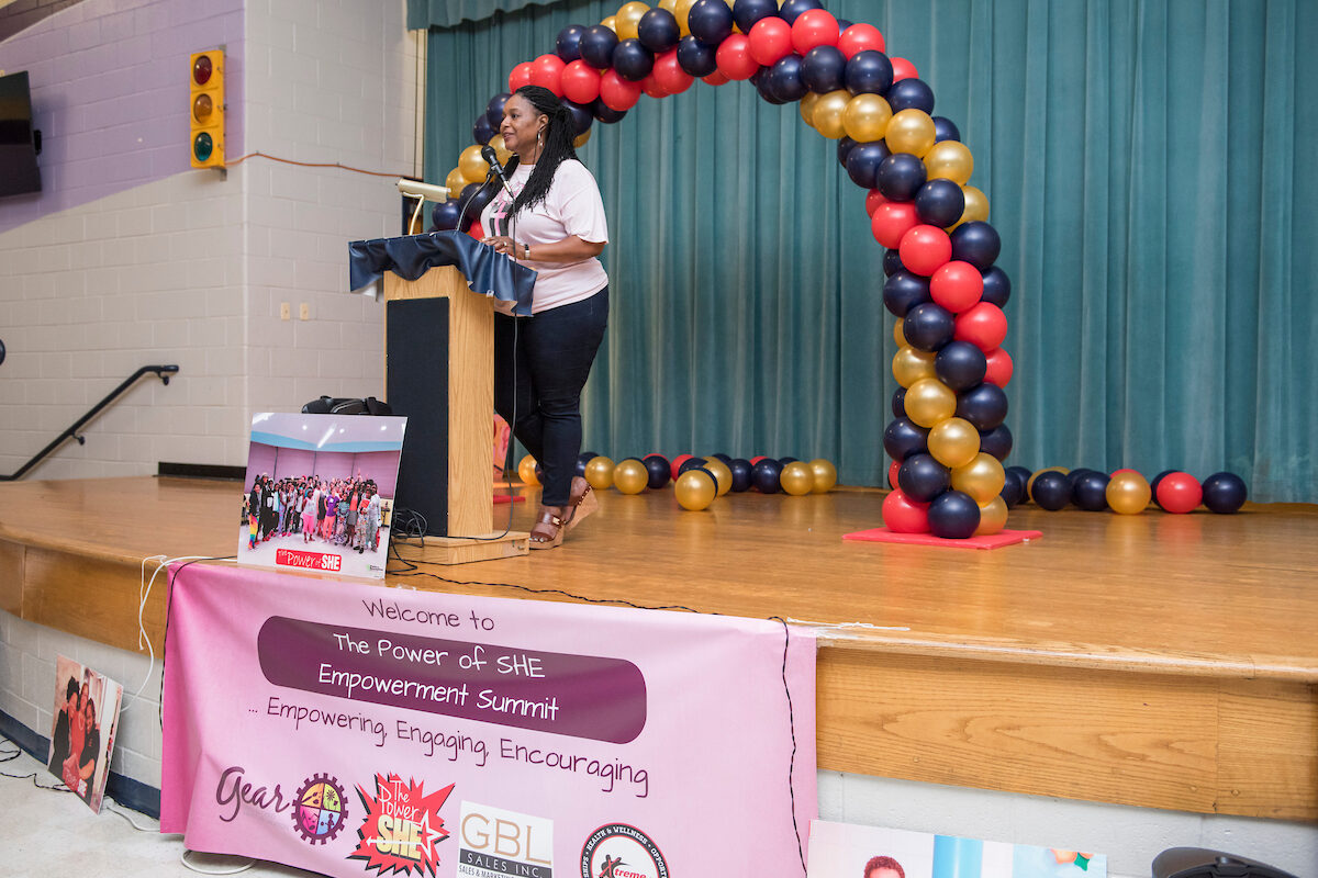 A woman stands behind a podium on a stage, addressing an unseen crowd. Behind her on the stage is a colorful balloon arch. A pink banner reading "The Power of SHE Empowerment Summit" hangs from the apron of the stage.