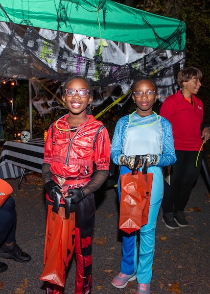 Two kids in Halloween costumes hold orange trick-or-treat bags. Behind them is a green tent decorated for Halloween with spiderweb garland and lights.
