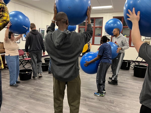 In a room with gray flooring, about eight adults hold large, blue exercise balls up over their heads.