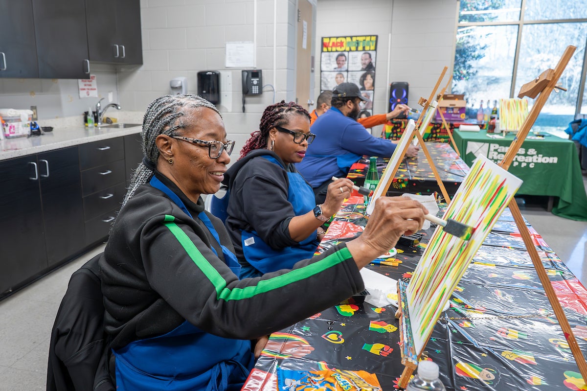 In a classroom with large windows, four adults sit at tables with plastic Black History-themed tablecloths, painting on canvases that sit on tabletop easels. They all wear blue aprons to protect their clothing.