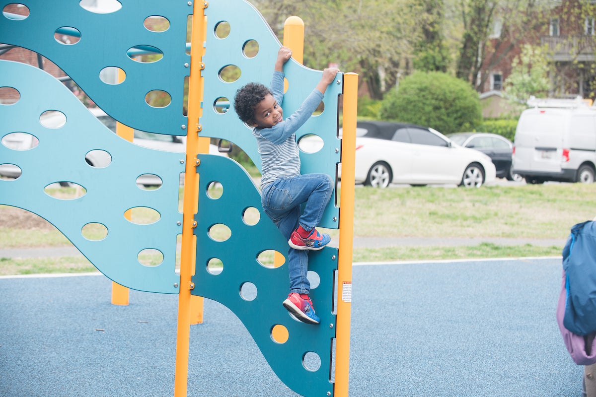 A child climbs on a vertical, blue climbing panel at an outdoor playground. The panel is wavy and filled with holes like Swiss cheese, for hand and foot-holds.