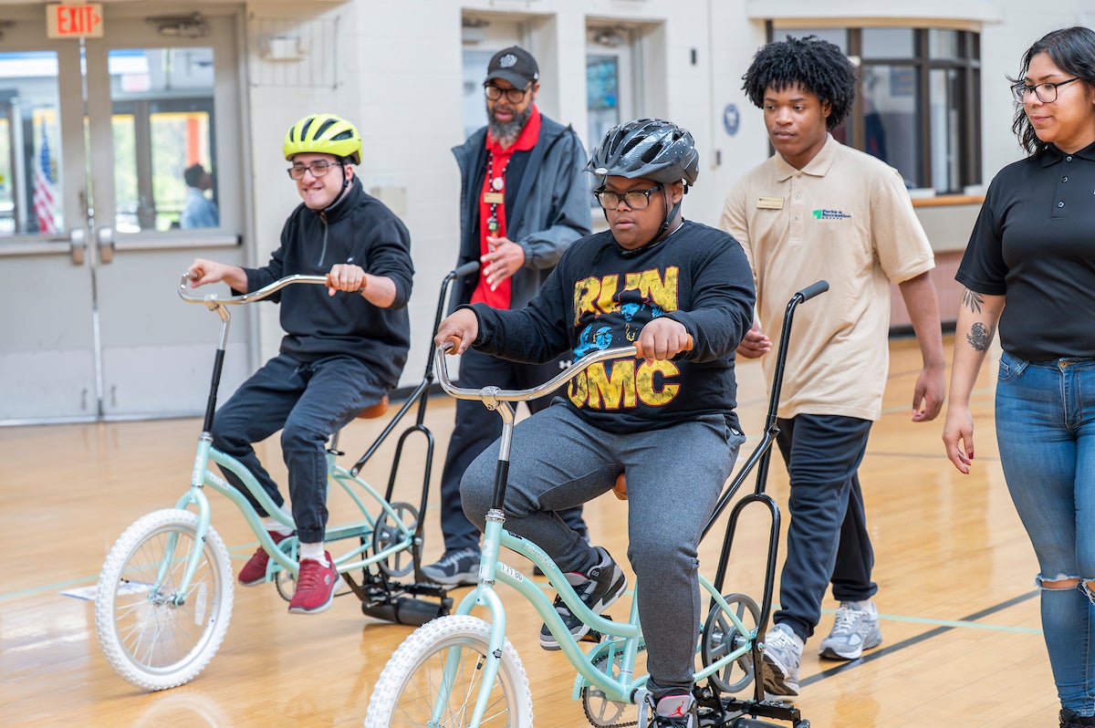 In a gymnasium, three staff members walk along behind two youths riding adaptive bicycles. The bicycles are mint green with white front tires.