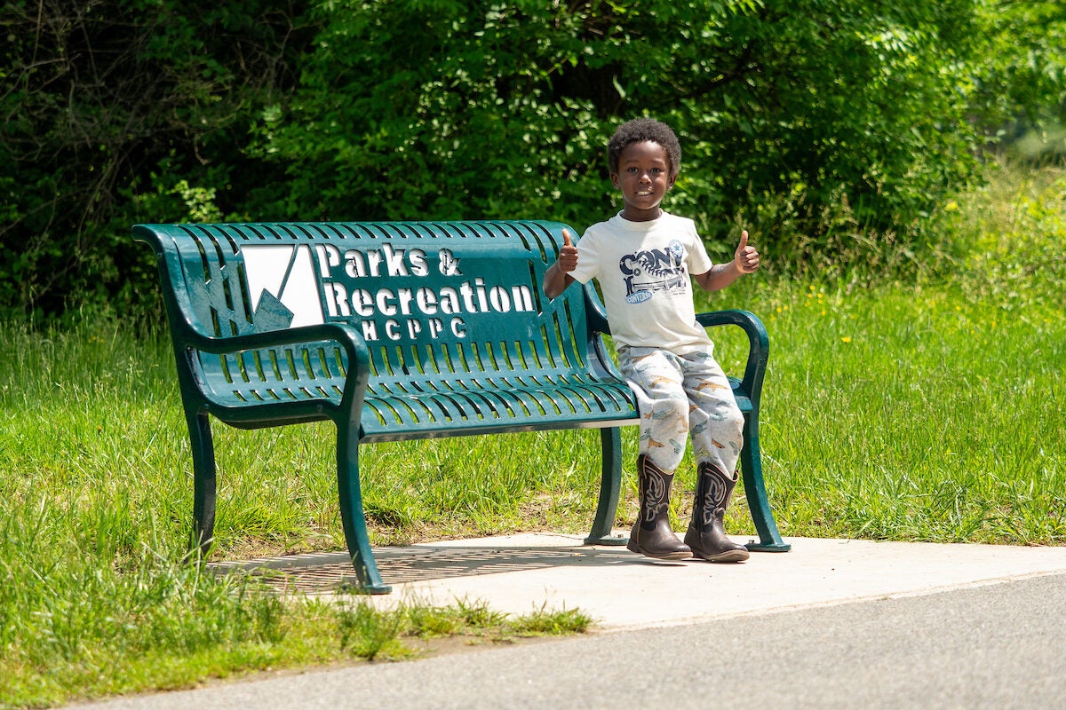 A small child sits on the far end of a green metal park bench with the words Parks & Recreation on it. The child is smiling and giving two thumbs up to the photographer. The bench sits on a concrete pad adjoining an asphalt walking path. Grass surrounds the concrete pad, with green brush and trees in the background.