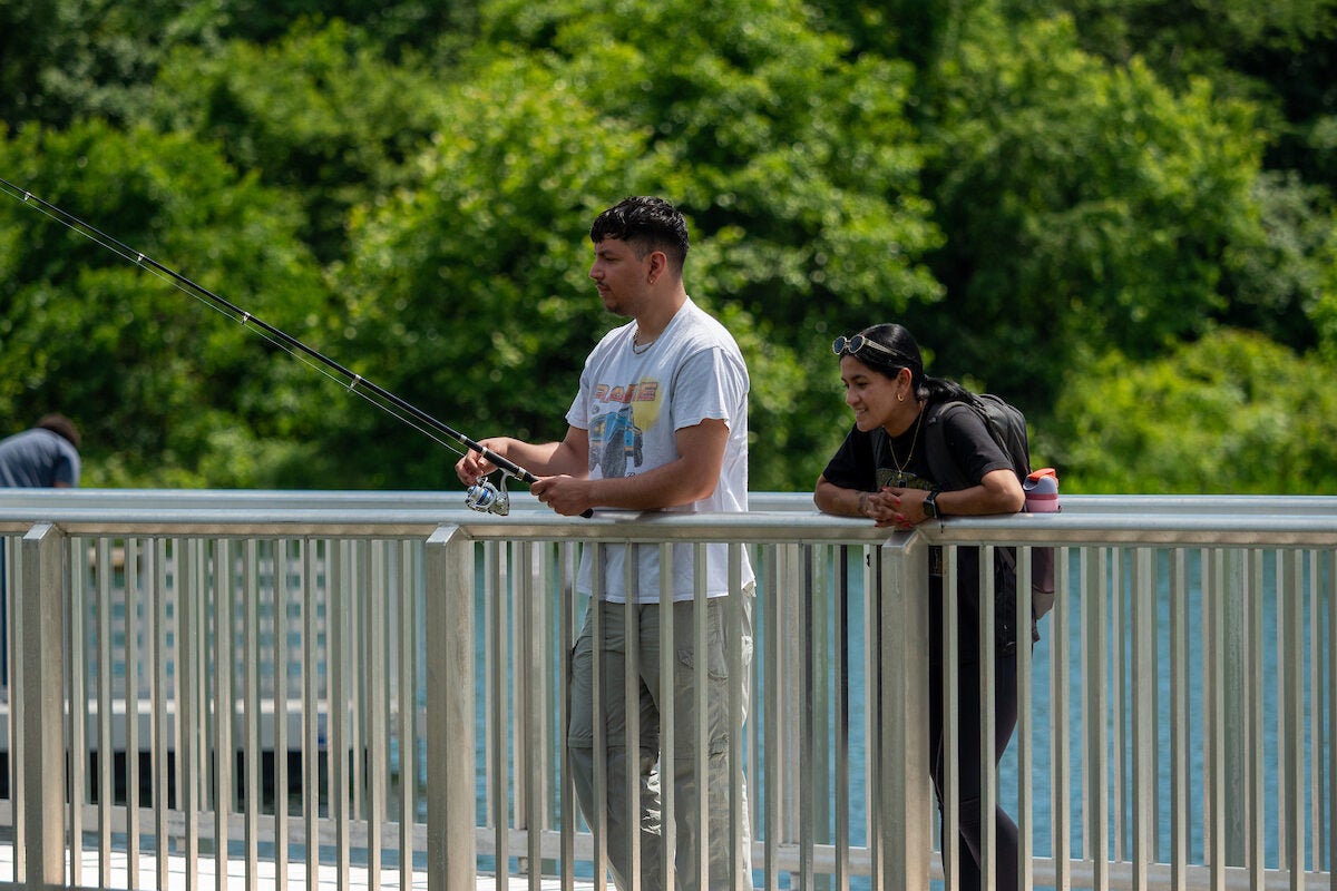 A man fishes off the side of a pier, while a woman at his side watches. It's a sunny day, and the water reflects the blue sky.