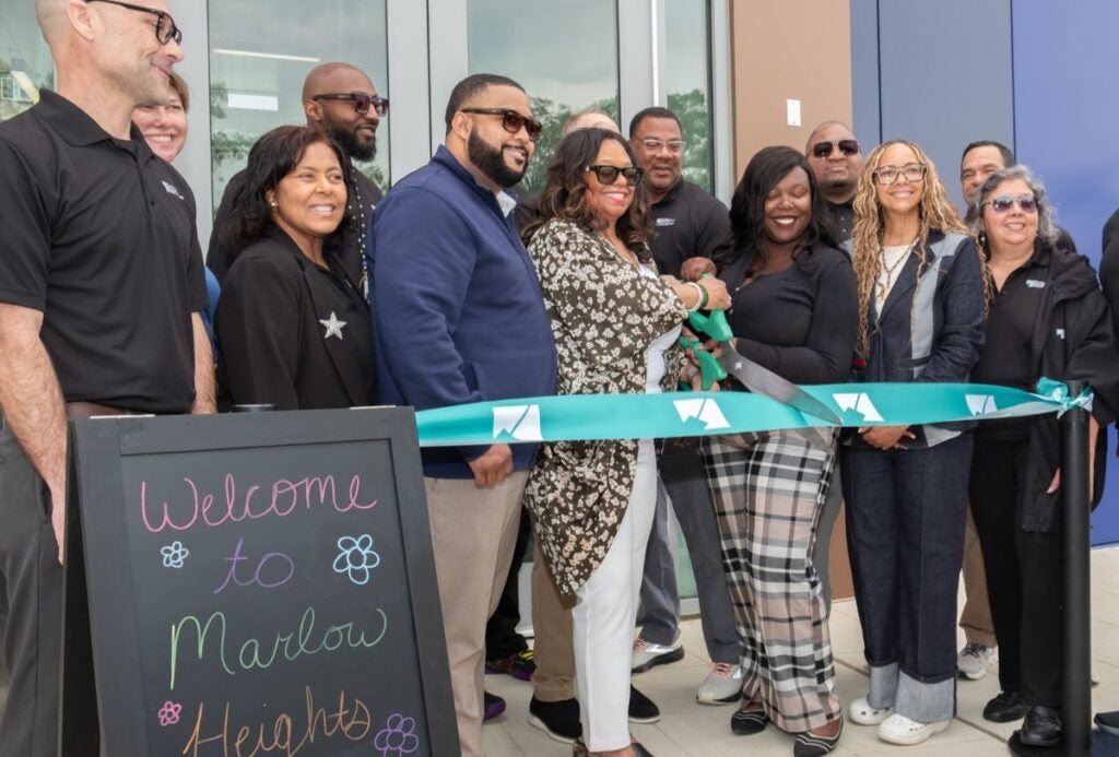 A dozen smiling adults, many wearing black Parks and Rec staff shirts, stand together in front of a building entrance, behind a ceremonial teal ribbon attached to two stanchions. Two women in the front row hold a giant pair of scissors, poised to cut the ribbon. A sidewalk sandwich board on the left reads, "Welcome to Marlow Heights..."