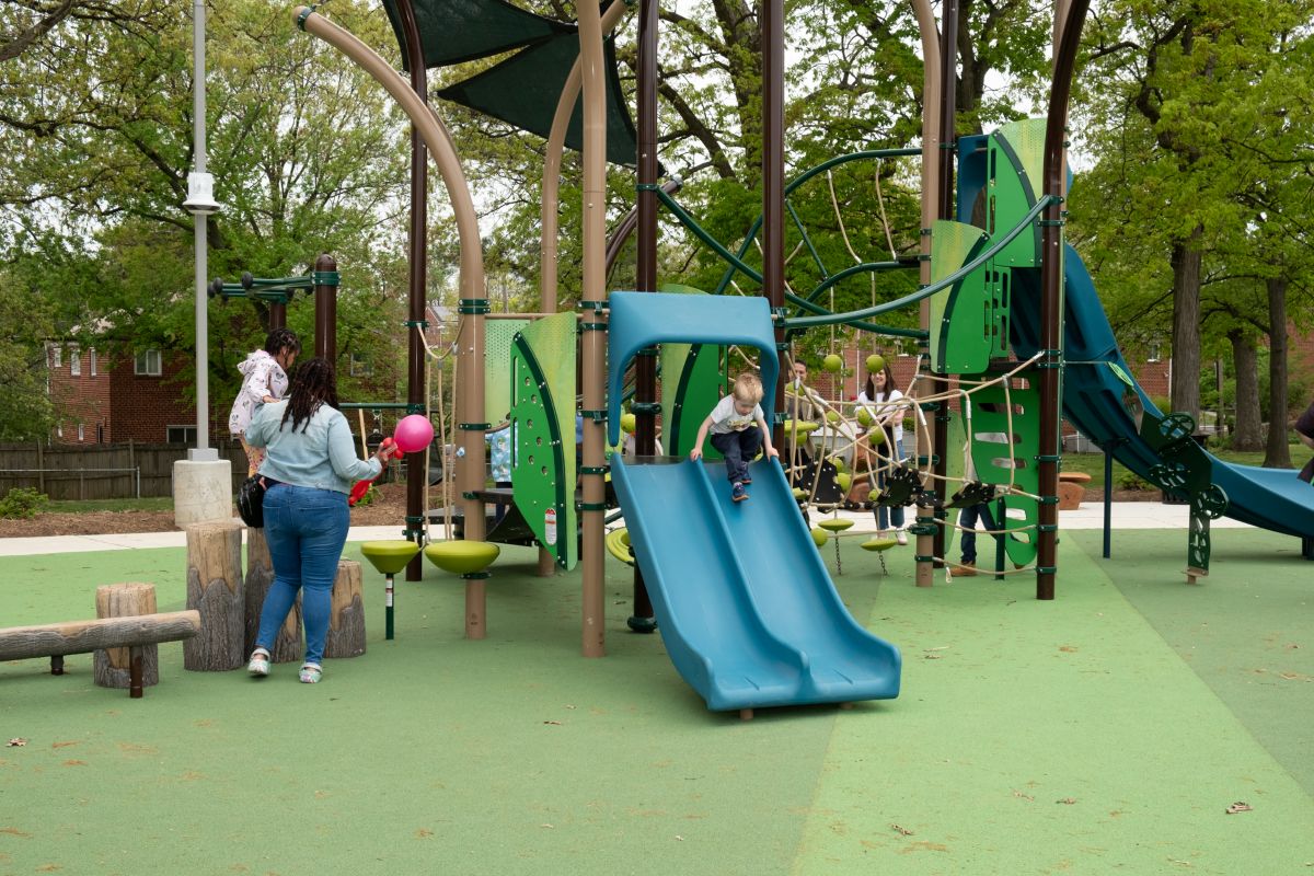 Kids play on a large, outdoor playground with multiple slides, climbing features, and platforms. The playground equipment is in shades of green, blue, and brown.