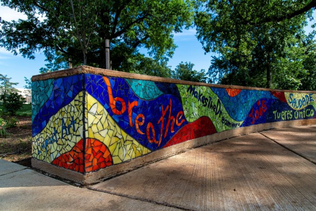 Close up view of one end of a long, curved, outdoor bench made of cement with a colorful glass mosaic inlaid on all sides. The mosaic, in shades of blue, green, yellow, and red, includes wave forms and words and phrases, including "empact art," "breathe," and more. Behind the bench are several tall trees.