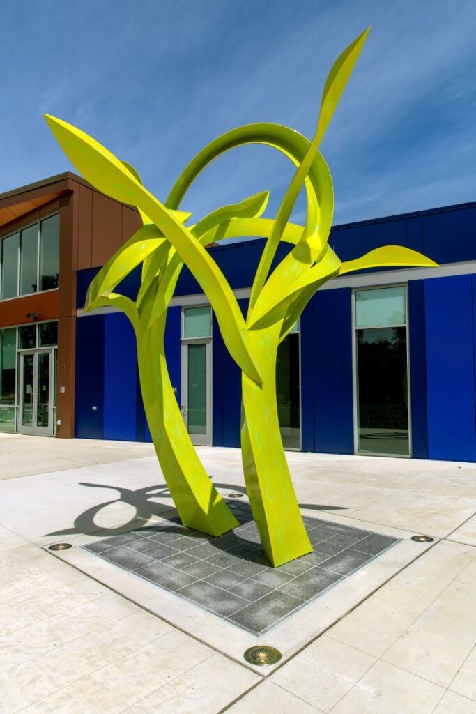 A large, neon green sculpture installed in a granite square in the concrete patio outside a large, blue and brown modern building. The sculpture swoops in a biomorphic form with several protrusions; overall it somewhat resembles curving blades of grass or plant seedlings.