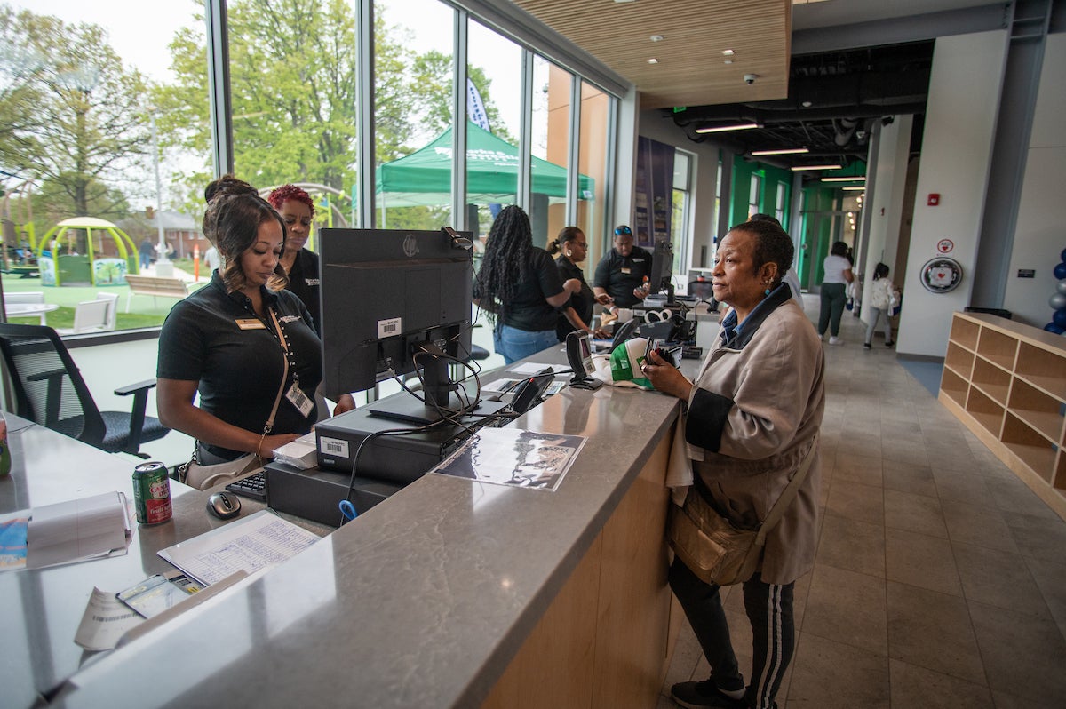 A woman stands at the front desk counter in a spacious lobby area with a wall of glass windows. Multiple staff members are behind the desk, some working on computers. Through the windows, we see a bench, a cafe-style table, and a large play structure.