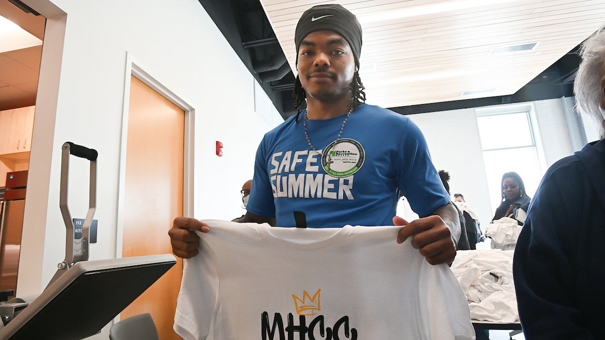 In a brightly-lit room, a young man wearing a blue SAFE SUMMER Parks and Rec shirt holds up a freshly-printed white t-shirt with the letters MHCC on it in a graffiti-style font. Behind him is a table piled with white t-shirts, and next to him is a press for printing the shirts.