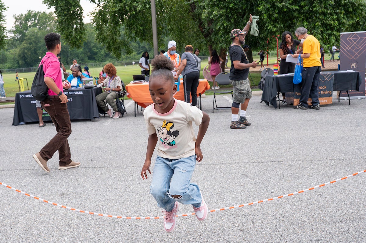 A girl wearing a Minnie Mouse t-shirt jumps rope in a parking lot. Behind her, where the parking lot meets a grassy field, are vendors and information tables.