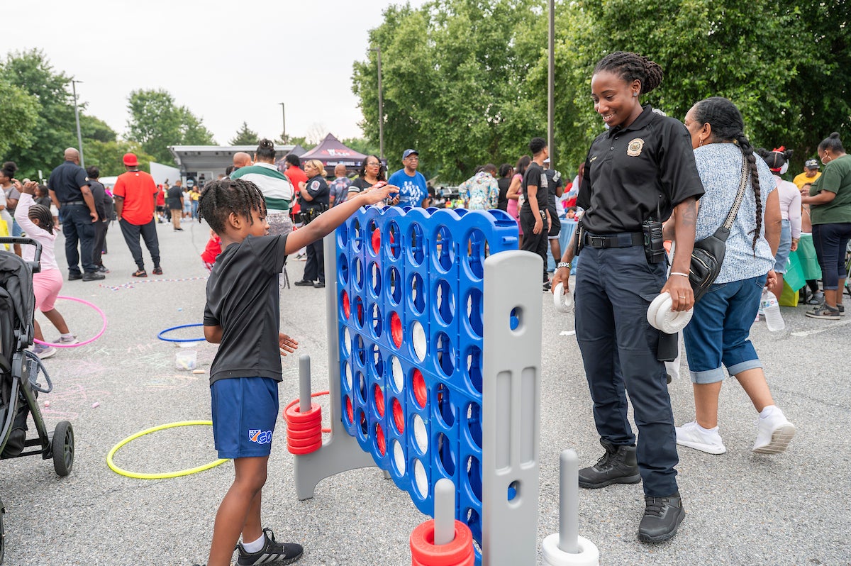 In a parking lot, a boy plays oversized Connect Four against a woman wearing a park police uniform and an amused expression. Behind them, kids play with hula hoops and jump ropes. There are adults crowded along the edge of the parking lot, where long information tables and vendors are set up. At the far end of the parking lot is a temporary bandshell.