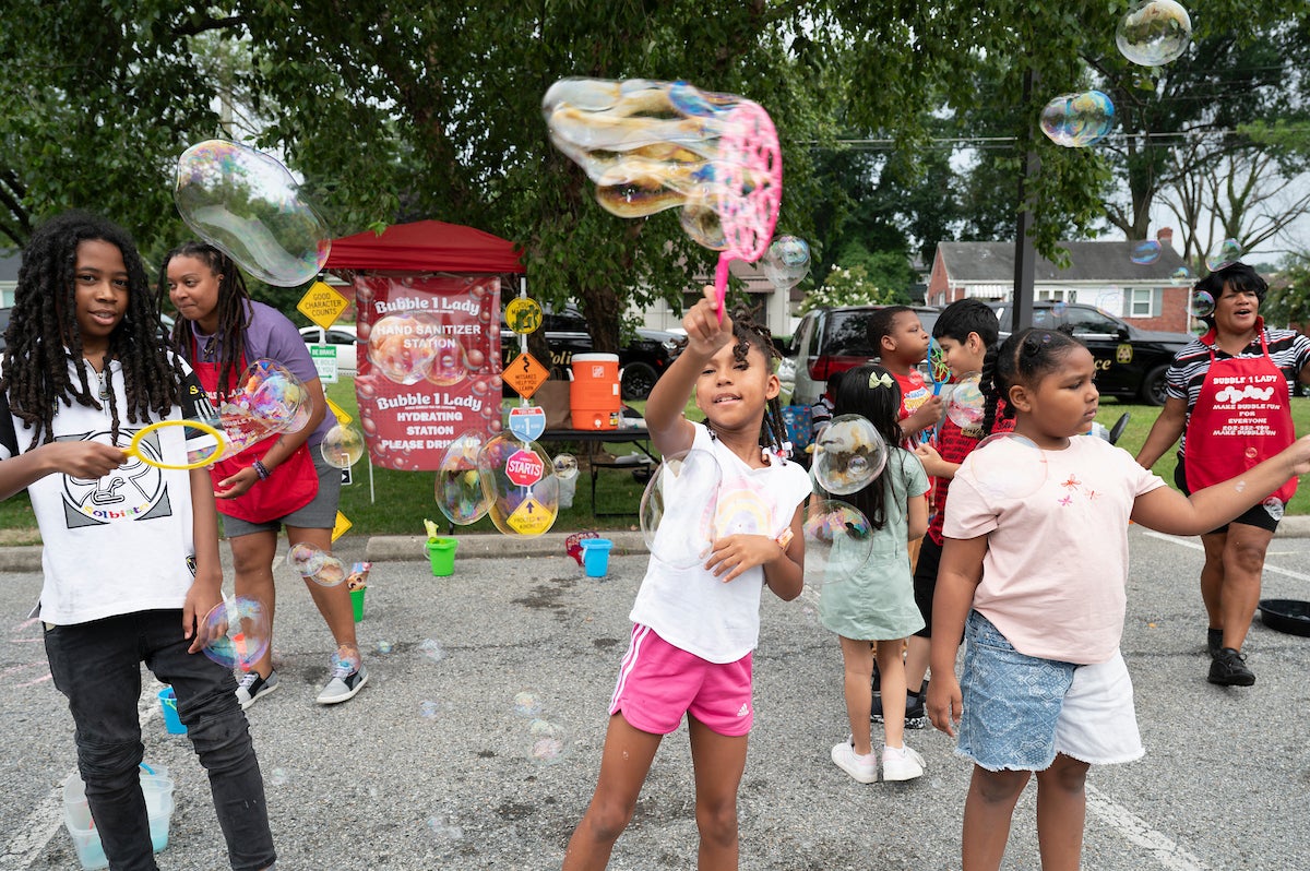 Half a dozen children use bubble wands of various sizes to create soap bubbles in an asphalt parking lot, at a station with a sign that reads Bubble Lady. Two women in red Bubble Lady aprons are assisting.