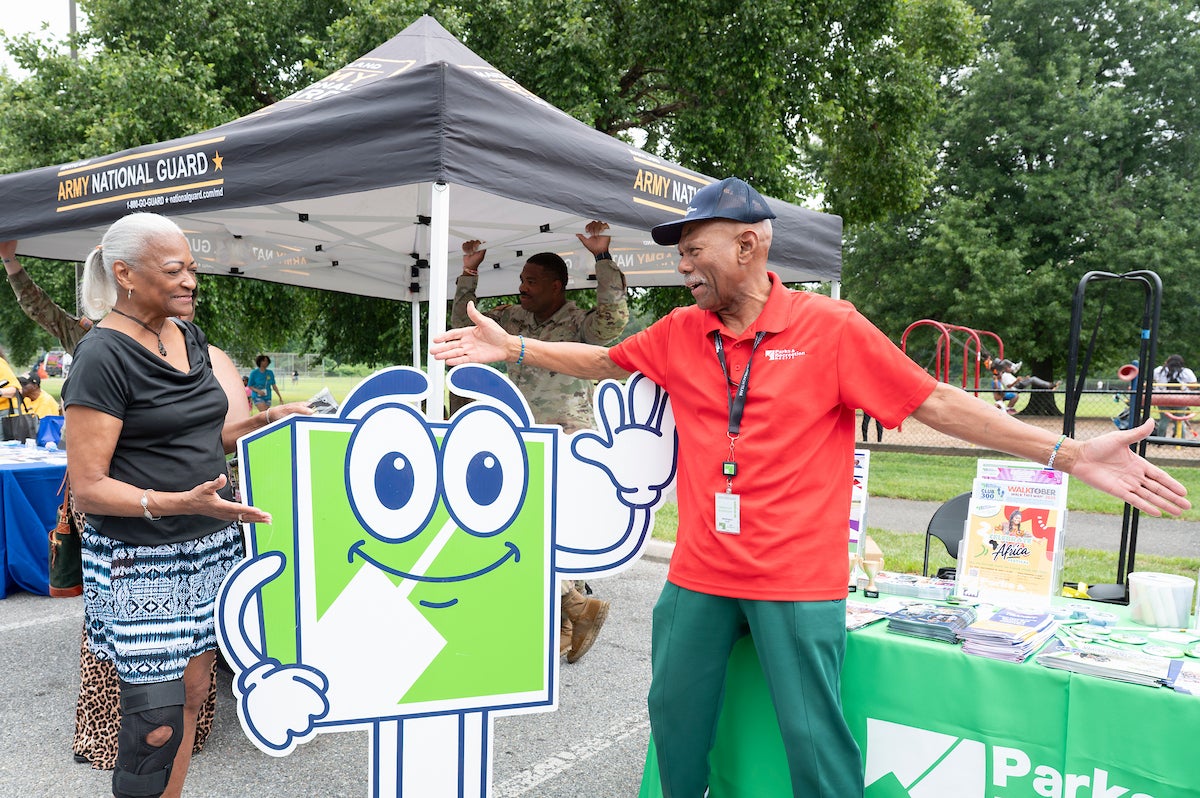 A man in a red staff polo shirt stands with arms outstretched talking to a woman next to a large cut-out of the green Parks and Rec mascot, FUN. Behind the man is an information table covered with flyers and program guides. In the background, through the trees, kids play on a playground.