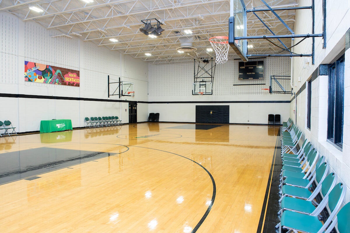 A gymnasium with a shiny wooden floor, multiple basketball hoops, and rows of folding chairs along the side walls. A scoreboard is mounted high up on the far wall.