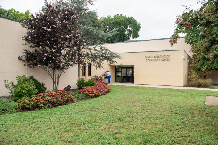 Exterior view of the entrance to a large, beige, cinderblock building with green trim. Letters on the building spell out NORTH BRENTWOOD COMMUNITY CENTER. There is a sidewalk leading to the entrance doors, next to a lush, green yard with landscaped flowerbeds and several flowering crepe myrtle trees.