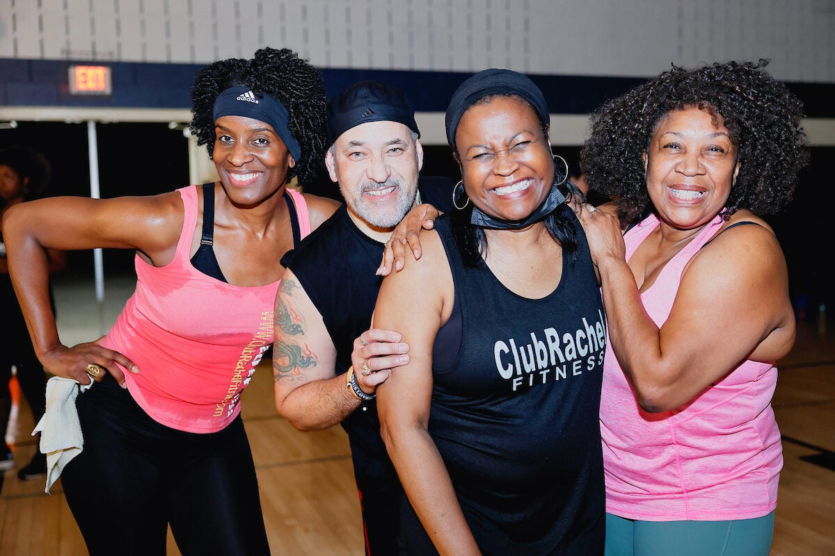 Four smiling adults in athletic clothing pose together in a gymnasium. One wears a black tank top with the words CLUB RACHEL FITNESS. They all appear to have just finished a taxing workout, with sweat marks on their clothing.