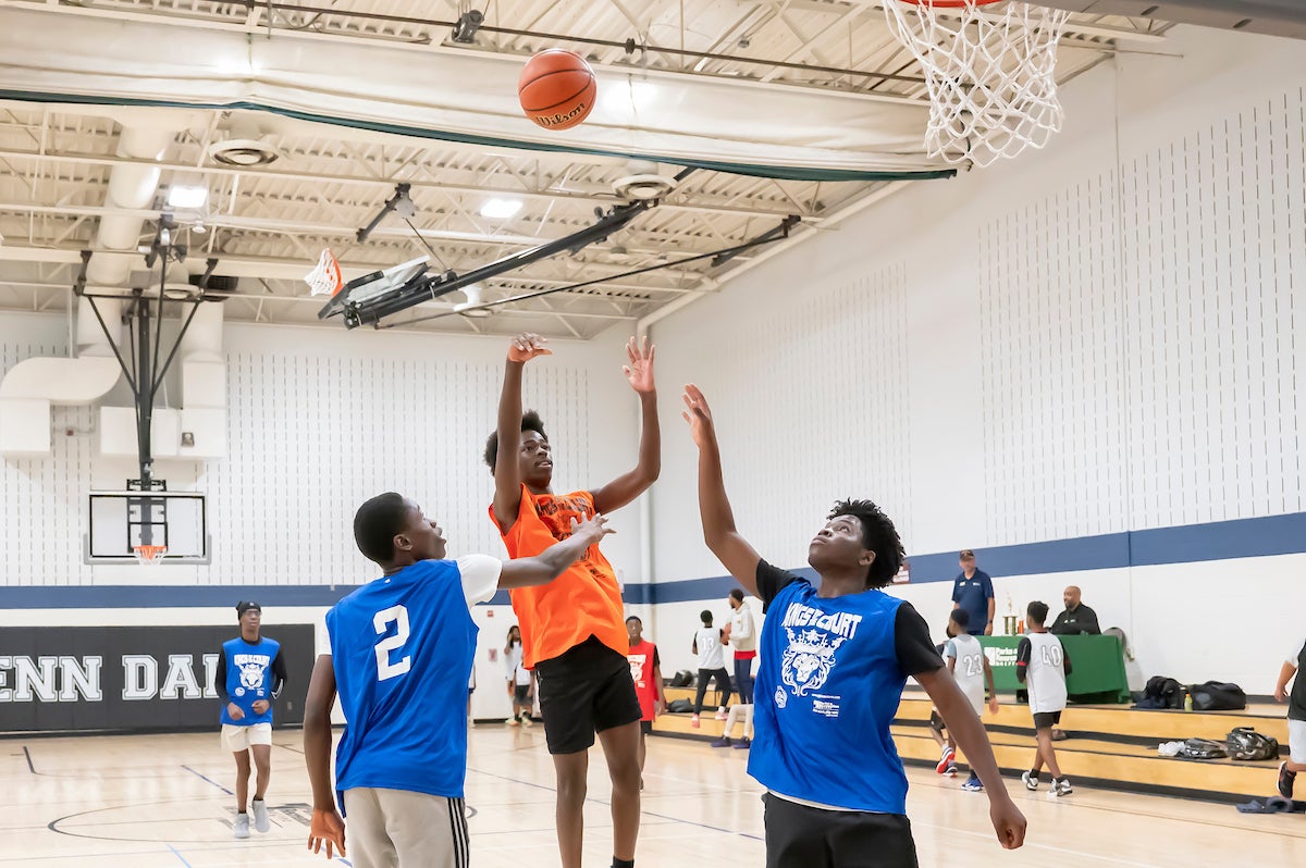 In a large gymnasium with the words GLENN DALE painted on the far wall, four teenage boys play basketball. One is in midair, having just thrown the basketball toward the hoop. Behind them to the right are about a dozen people on low, wooden bleachers/benches, and a staff member sitting at a table.