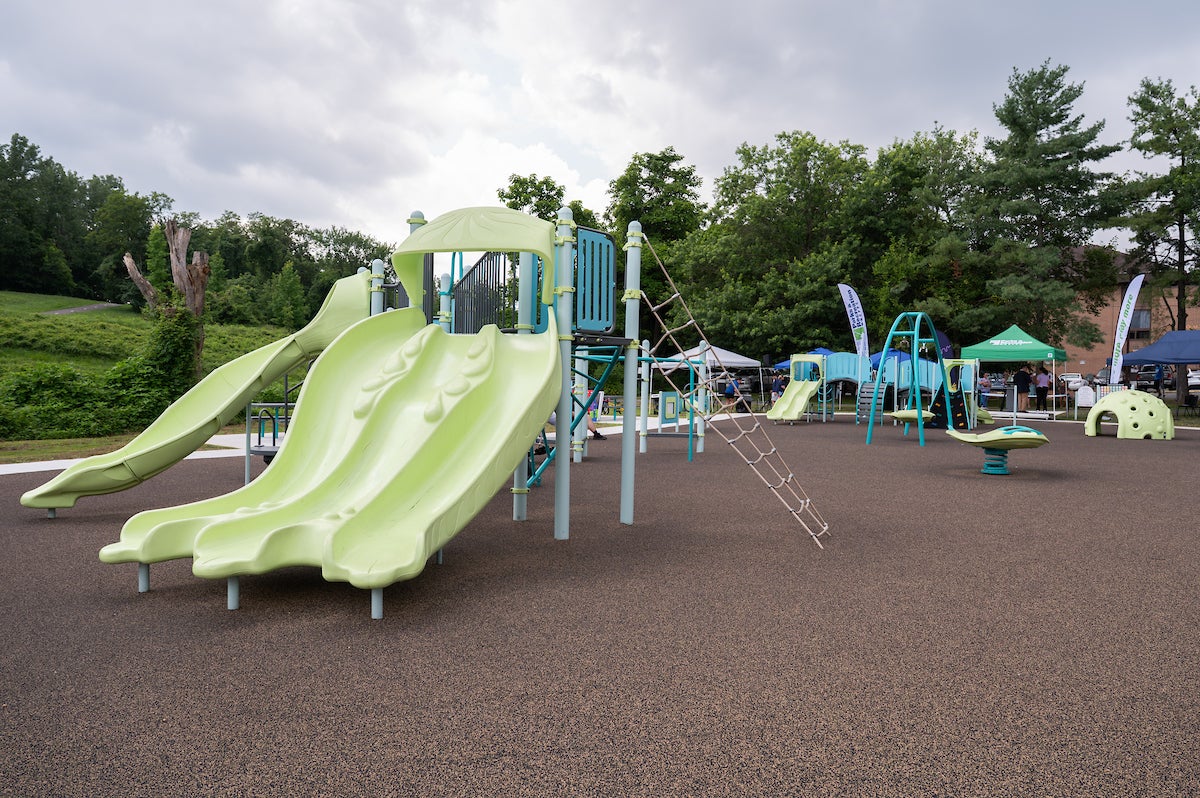 An outdoor playground with multiple play structures, including swings, slides, rope webs, monkey bars, platforms, rocking elements, and spinning elements. Beyond the playground are grass and trees.