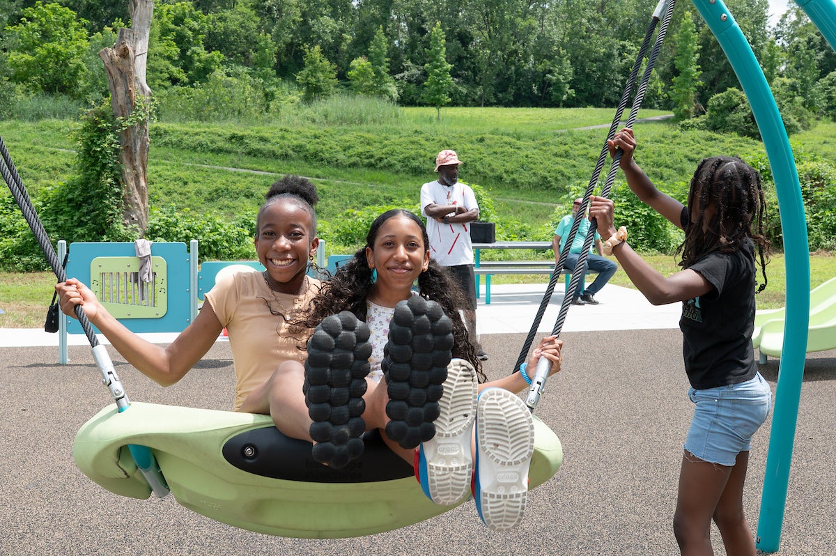 At an outdoor playground, two youths swing on a large, plastic, dish-style swing while a third holds onto the rope to help push them. In the background are some musical instrument play elements and a picnic table.