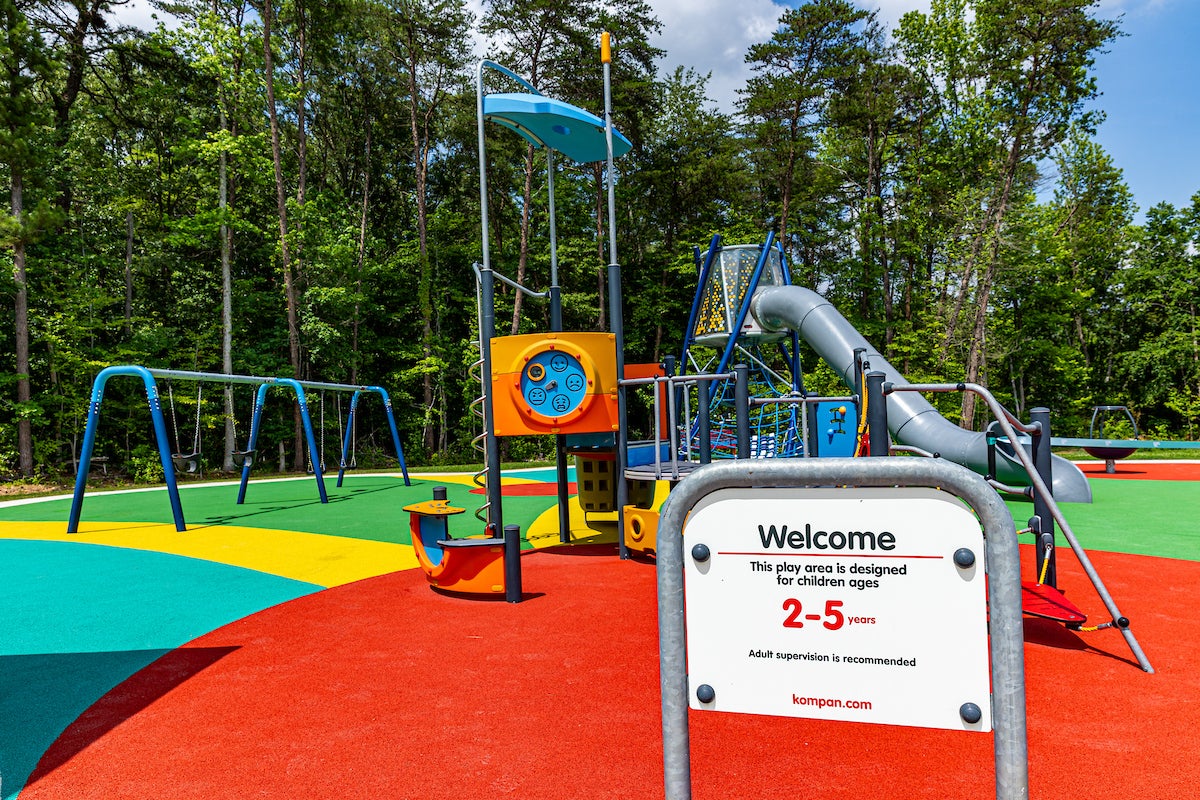 A colorful outdoor playground featuring swings, slides, platforms, games, rope webs, spinning elements, and more. A sign posted near the edge reads WELCOME, THIS PLAY AREA IS DESIGNED FOR CHILDREN AGES 2-5 YEARS.