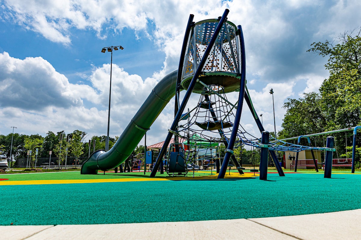 A tall play structure on an outdoor playground featuring a tube slide down from a high platform encased in plexiglass walls, and a rope web below the platform as the only visible means for climbing up.