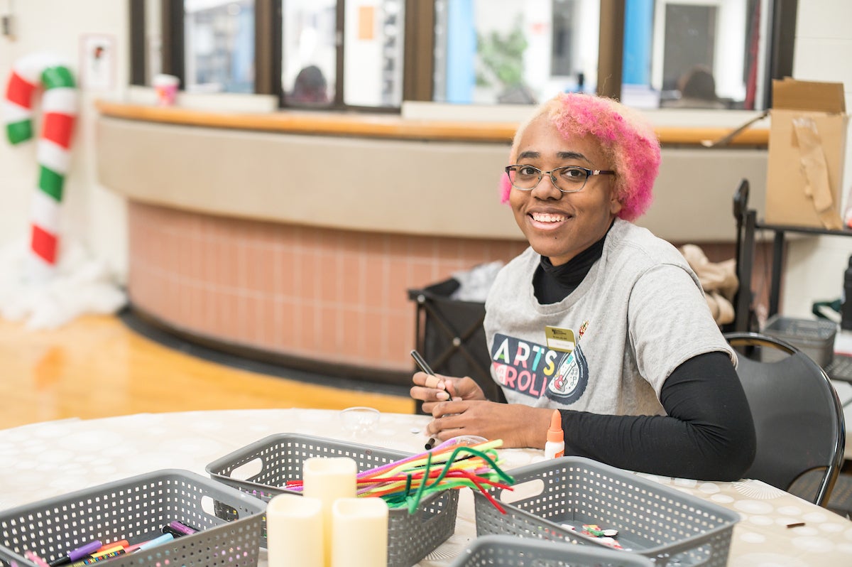 A smiling woman with pink hair wearing an Arts on a Roll t-shirt sits at a round table with craft supplies arrayed out in front of her, working on a holiday ornament. Behind her is a reception window counter, and a large candy cane decoration.