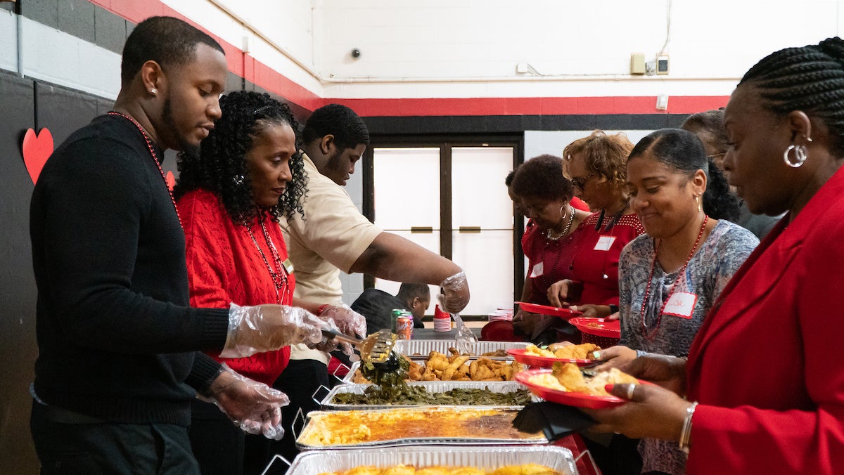 In a gymnasium decorated for Valentine's Day, three staff members stand behind a long buffet table, serving food to adults holding red plastic plates. Most of them are wearing red for the Valentine's theme.