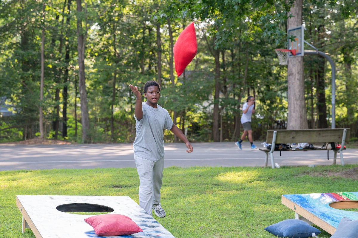 A child tosses a large, red beanbag toward an oversized game of cornhole on the grass. Behind him is an outdoor basketball court surrounded by tall trees.