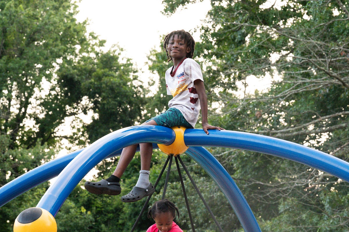 A smiling child sits atop a large swing support structure on an outdoor playground. Below him, a child in pink plays on a swing suspended from four ropes.