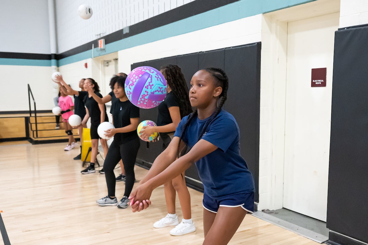 In a gymnasium, seven teen girls stand near the padded wall in a line, each with a volleyball. The girl closest to the camera has her arms out and hands laced together while practicing hitting her ball in the air, with a concentrated look on her face.