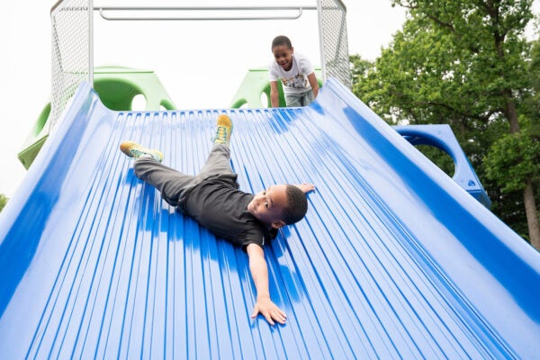 A boy slides sideways down an extra-wide, blue plastic slide at an outdoor playground, while another boy gleefully watches from the platform at the top of the slide.