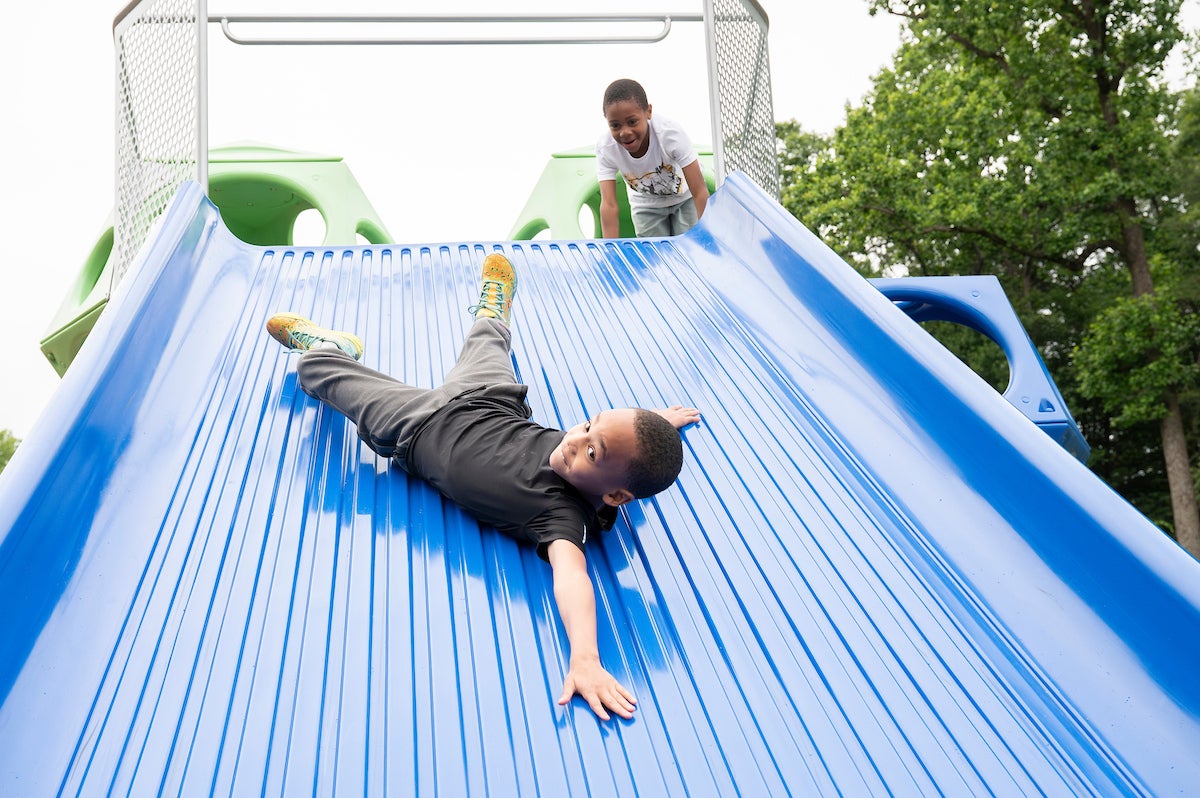 A boy slides sideways down an extra-wide, blue plastic slide at an outdoor playground, while another boy gleefully watches from the platform at the top of the slide.