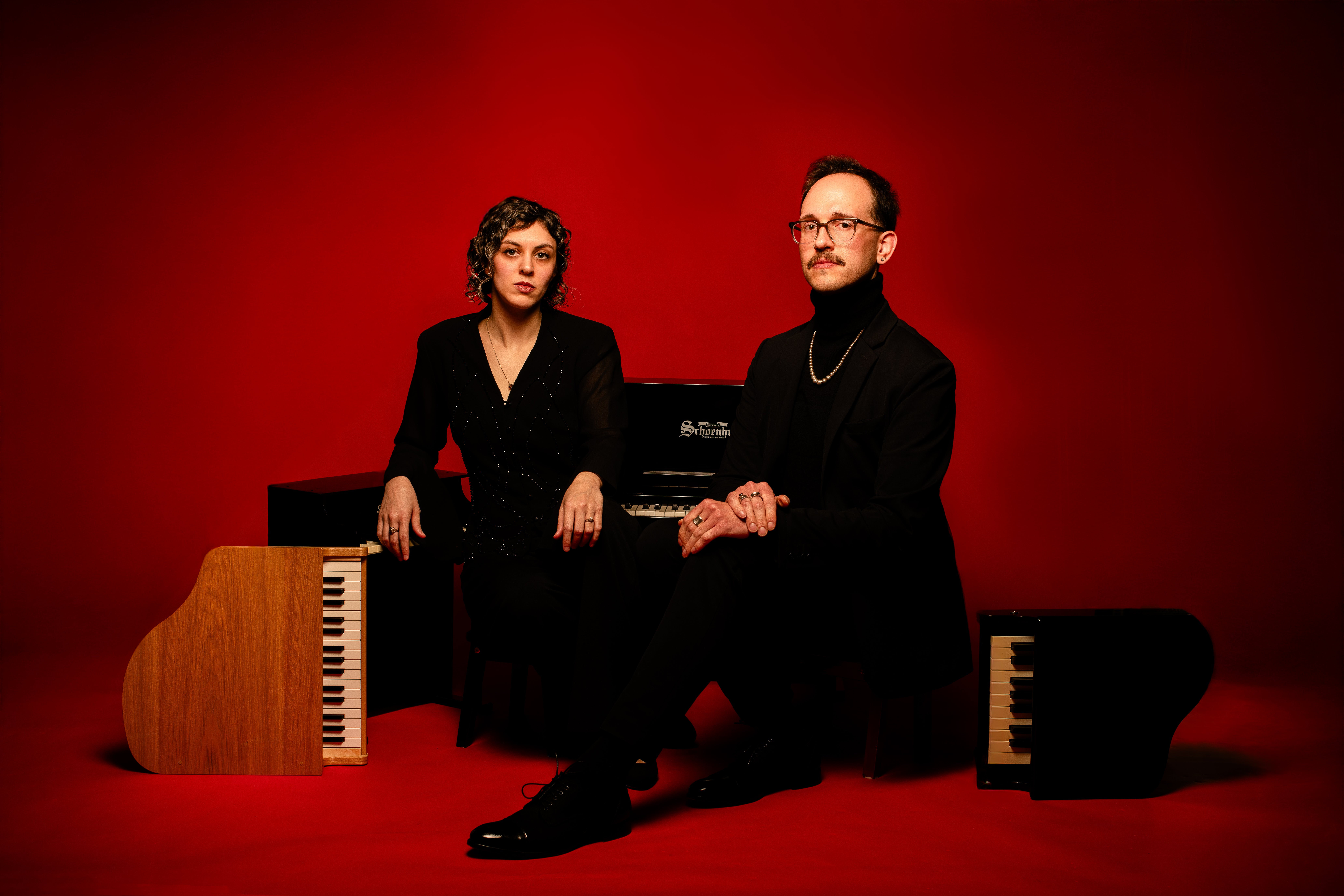 Studio portrait of two people in black attire seated at keyboard instruments against a solid red background.