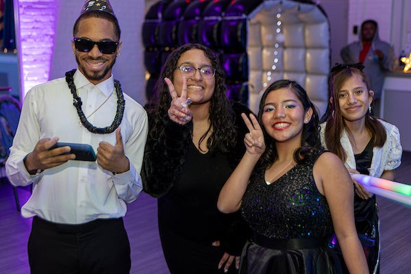 Four smiling teens pose for a photo in formal attire, giving peace signs and thumbs up. One boy wears sunglasses, a novelty party necklace, and a party hat. Behind them, the room is dimly lit with purple LED party lights.
