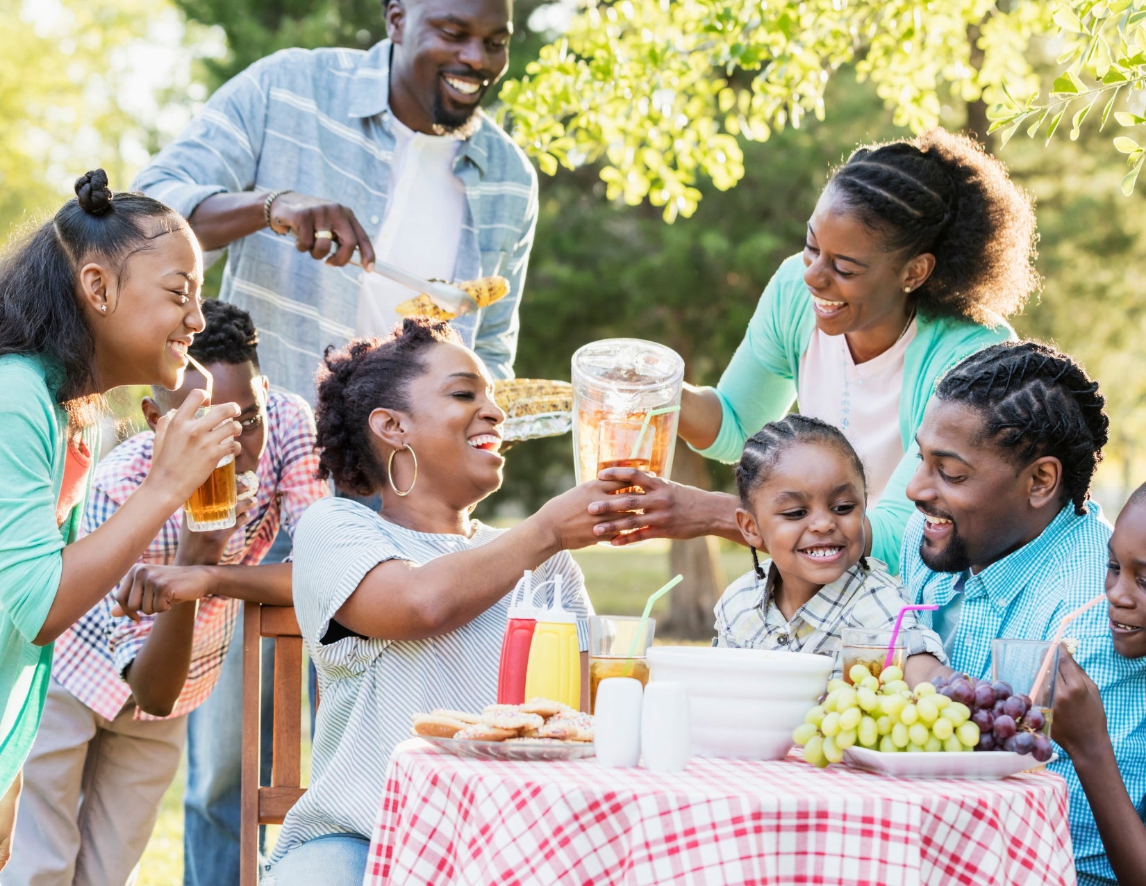 Group of people sharing food and drinks at an outdoor picnic table, with grilled corn, fruit, and condiments.