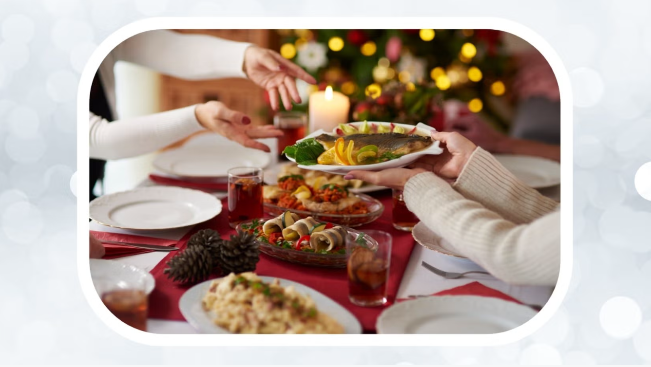 A close-up view of hands passing a plate with a fish on it across a dinner table that is laid with many plates and contains multiple dishes of food. Hands on the other side of the table reach for the fish dish. In the background is a blurry Christmas tree.