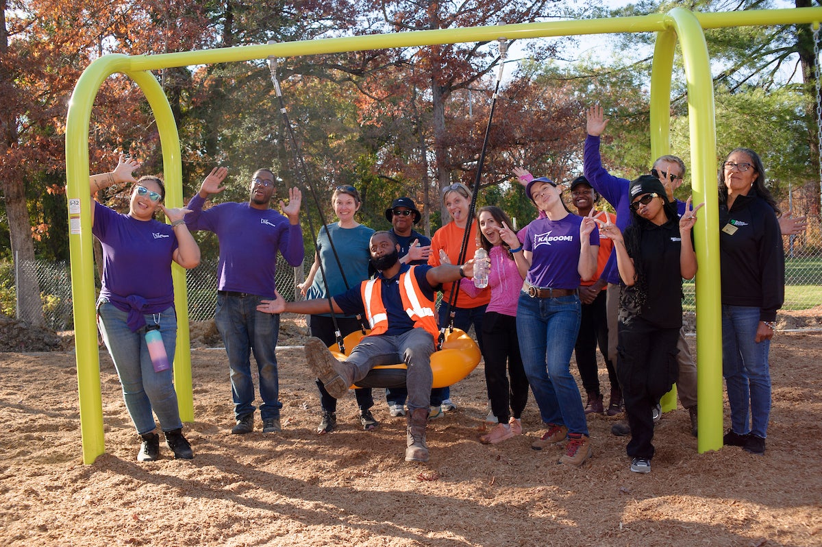 A dozen adults smile, wave, and pose for a photo on an outdoor playground. One man in an orange visibility vest sits in a large, orange, donut-style swing on a yellow swing set, with the other adults clustered around him on both sides.