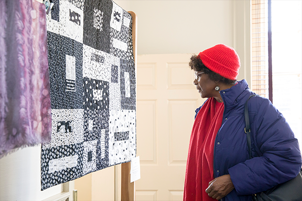 A woman wearing a red hat and a blue coat looking at a textile quilt at a display.