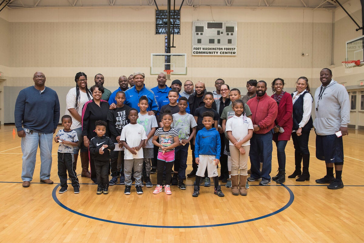 About 25 adults and a dozen kids pose for a photo in the middle of an indoor gymnasium. A scoreboard mounted high on the wall behind them reads FORT WASHINGTON FOREST COMMUNITY CENTER.