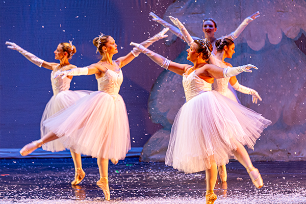 Five dancers in white costumes with long tulle skirts dance on a stage covered in confetti or flower petals.