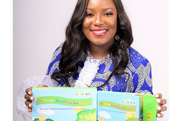 A smiling woman in a blue patterned dress displays an open children's book with the title "The Yoruba Sound Book for Children." Next to the pages on the right edge of the book is an electronic pad with a speaker and several buttons.