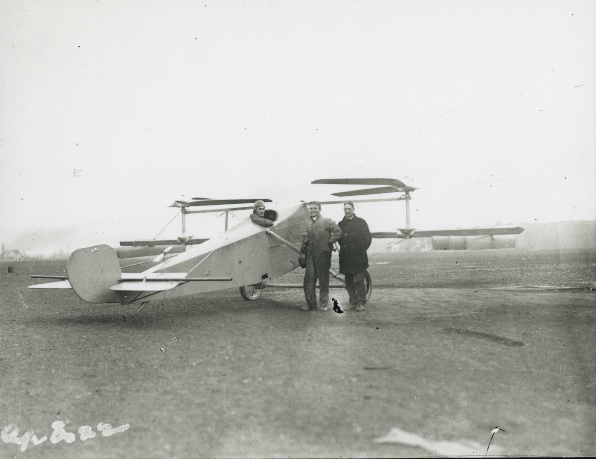 Historic, black-and-white image showing two men standing on a field next to a small aircraft with a pilot sticking out of a cockpit on the top. The body of the aircraft resembles the body of a biplane, but in place of wings, this aircraft has two horizontal propellers.