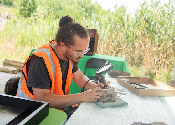 A man holding a rotary tool looking at a rock through a large magnifying glass.
