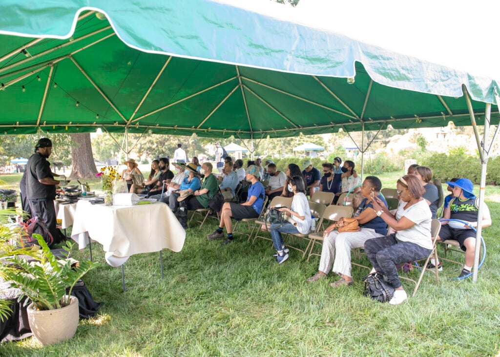 A group of people sitting in chairs under a large green tent watching a man behind a table cook