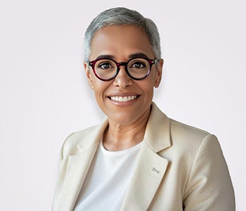 A black woman with short white hair smiling at the camera with a white background. she is wearing a white suit jacket and red glasses.