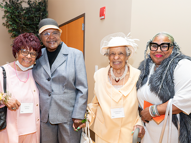 Four women standing side by side smiling. They are wearing light pink, gray, yellow, and white and gray outfits respectively. They are standing in a gymnasium that's filled with tables and chairs to celebrate people who are over 100 years old.