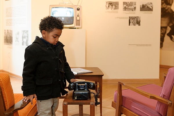 A child in a black coat standing in a interactive set piece of chairs, a table, and an old television. The set is designed to look like a 50s household, and has information regarding civil rights activists in America.