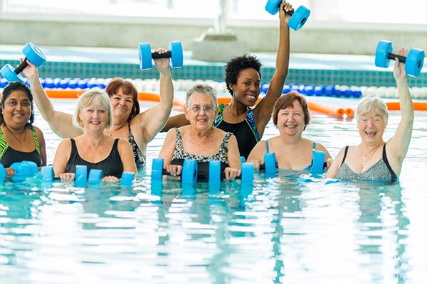 A class of women in an indoor pool, all smiling and holding blue hand weights.
