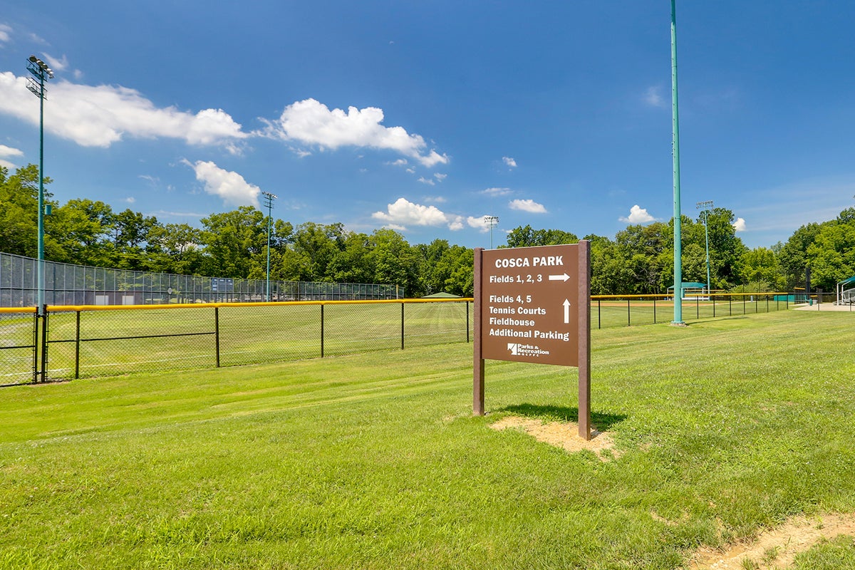 A brown sign that says COSCA PARK in the grass in front of an athletic field with directional arrows for Fields 1-3, Fields 4 & 5, Tennis Courts, Fieldhouse, and Additional Parking.